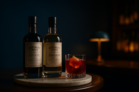 Row of Asterley Bros bottles including ESTATE English Sweet Vermouth and SCHOFIELD'S English Dry Vermouth, beside a crafted Negroni in a rocks glass